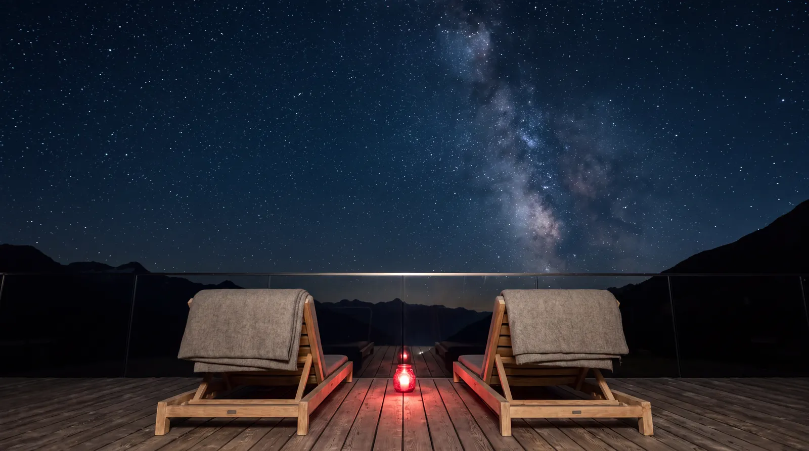 Die Terrasse bei Nacht: zwei Holzliegen mit Wolldecken, eine rote Laterne, darüber die Milchstraße.