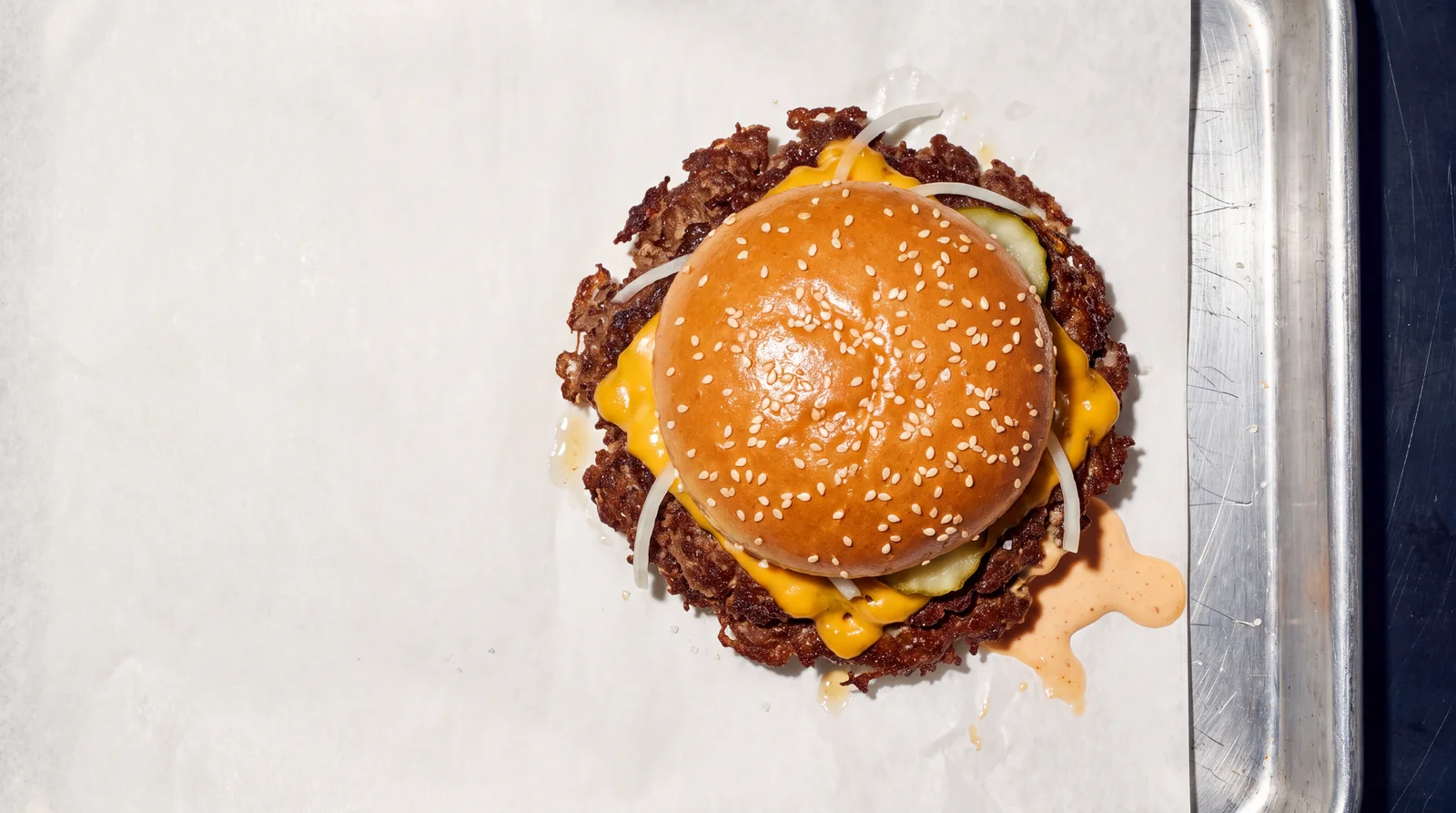 Ein Smash Burger von oben auf Papier und Stahltablett: Sesambrötchen, Käse läuft über die Spitzenkruste, Zwiebel, Gurke, Sauce.